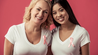 Women against breast cancer. Beautiful mature and Asian women with pink ribbons on their chests are looking at camera and smiling, on red background