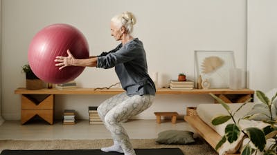 Side view of aged active woman in sportswear holding fitball in front of herself while doing squats on the floor of bedroom in the morning